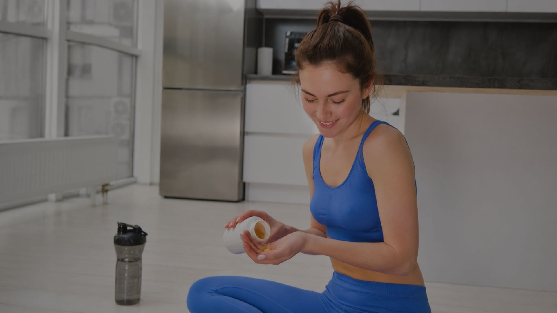 Woman in blue athletic wear sitting on the floor with a shaker bottle and container in a kitchen.