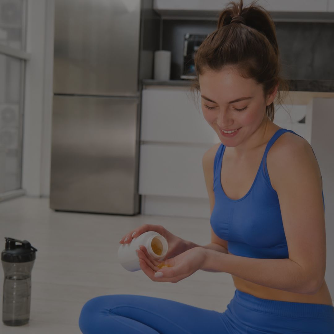 Woman in blue athletic wear sitting on the floor with a container and shaker bottle in a kitchen.